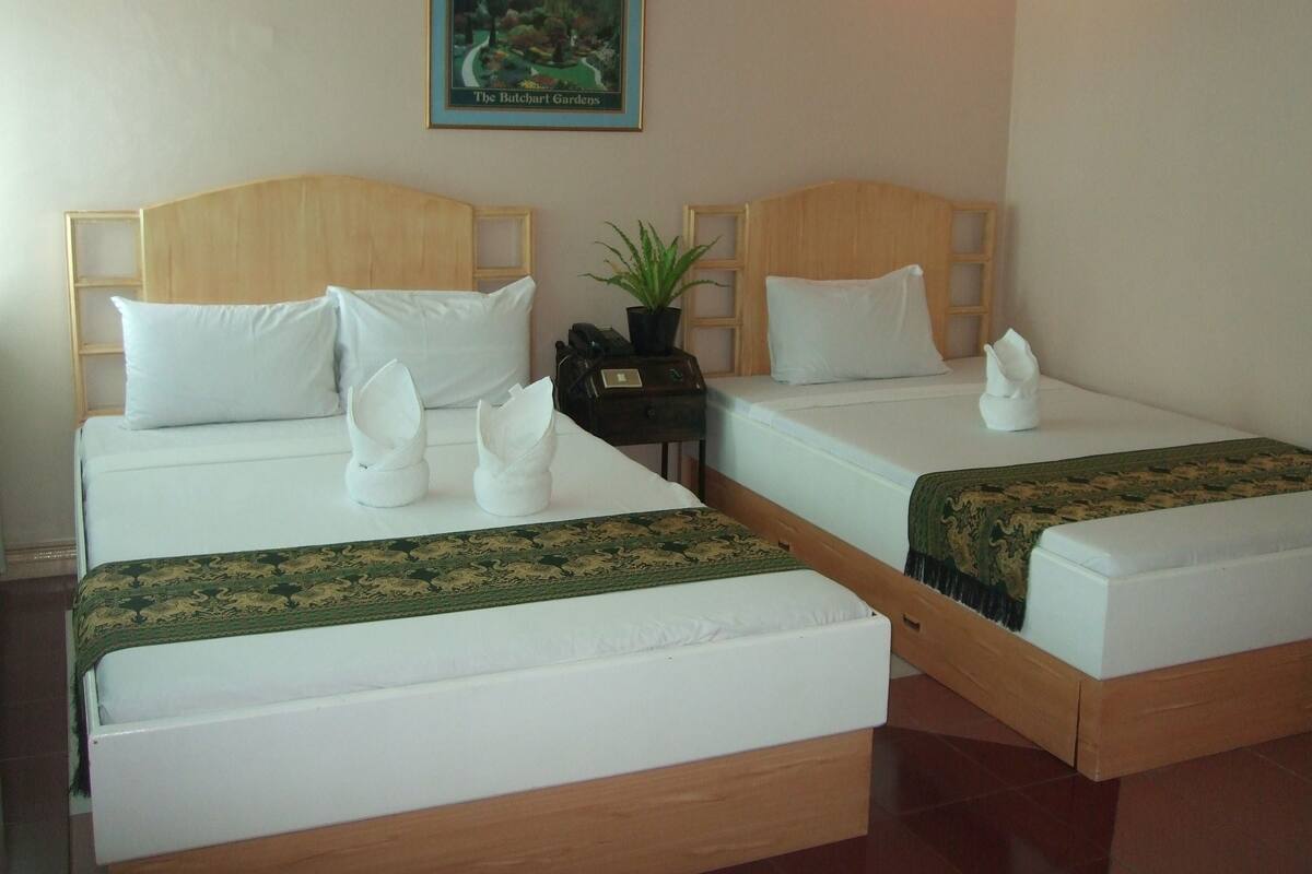 Two single beds with white linens, a framed picture of a garden on the wall, and a small potted plant on a nightstand between them.