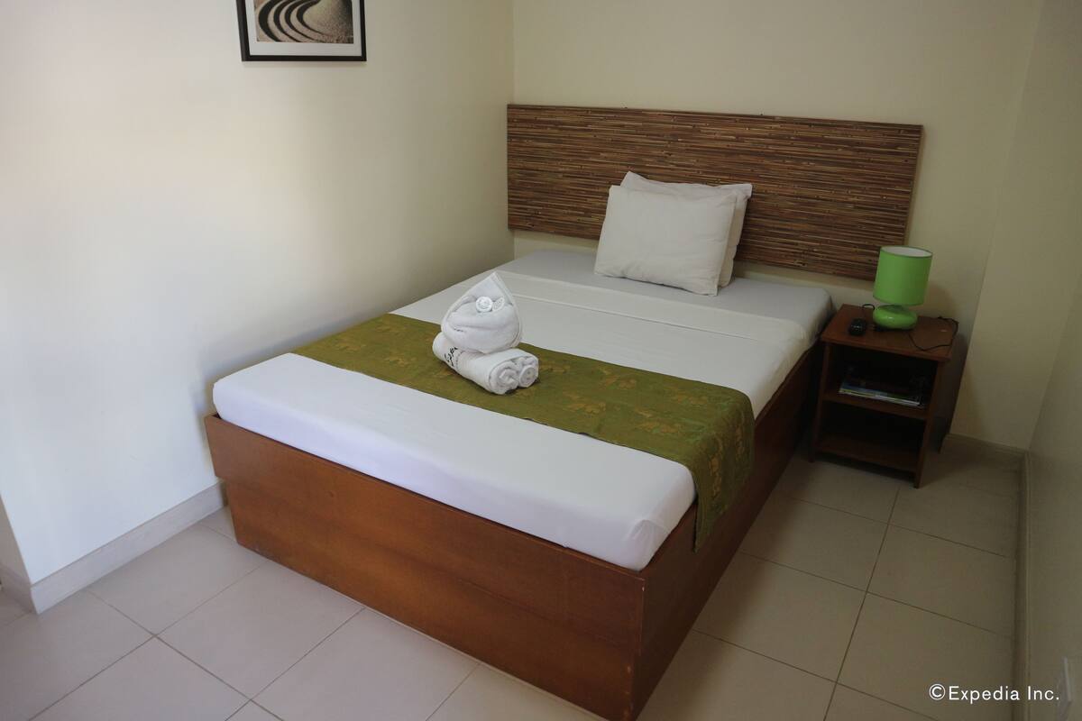 A neatly made bed with a wooden headboard, a green throw blanket, and a rolled towel on the footboard.