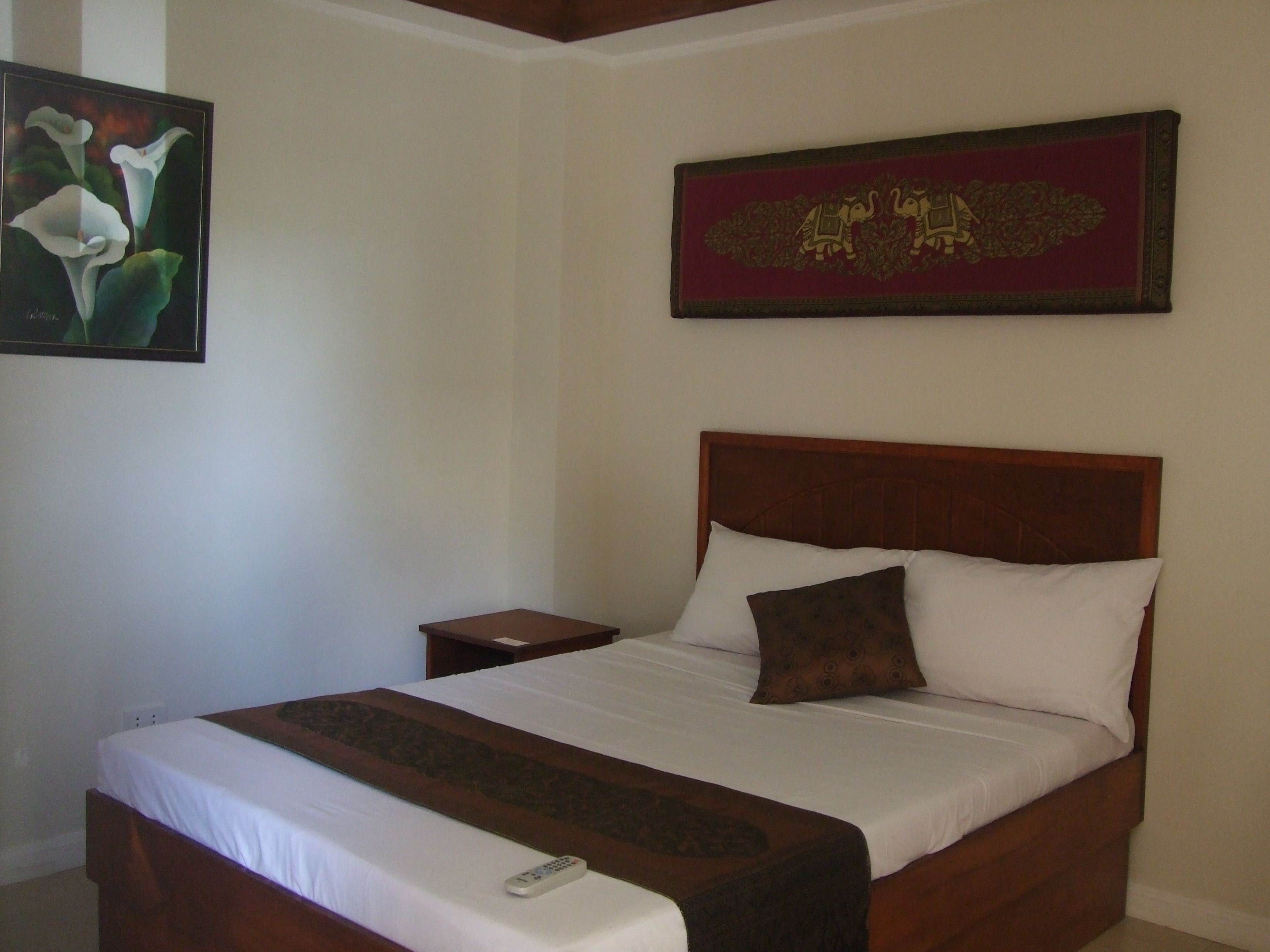 A neatly made bed with a remote control on the side, a nightstand, and a framed picture of flowers on the wall.