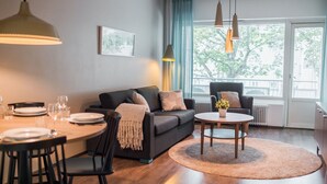 Una sala de estar moderna con un sofá, una mesa de comedor y sillas. Hay una ventana con cortinas, un radiador y una mesita con un florero.