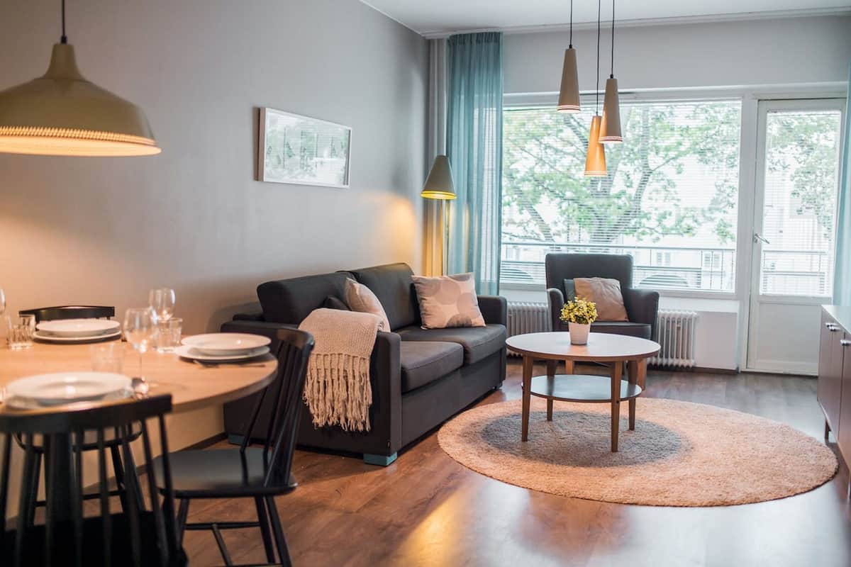 Una sala de estar moderna con un sofá, una mesa de comedor y sillas. Hay una ventana con cortinas, un radiador y una mesita con un florero.