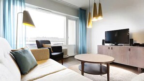 Un moderno cuarto de hotel con sofá, sillón, mesa redonda, televisor y vista a la ciudad a través de amplias ventanas.