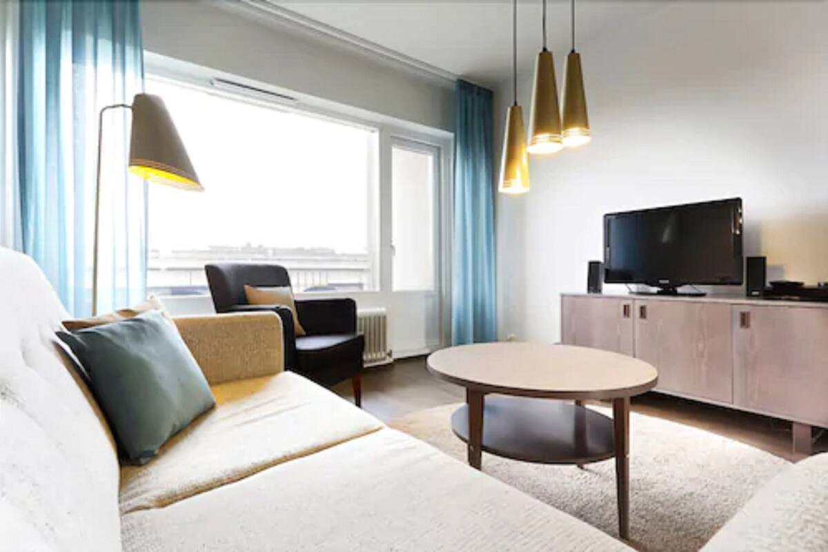 Un moderno cuarto de hotel con sofá, sillón, mesa redonda, televisor y vista a la ciudad a través de amplias ventanas.