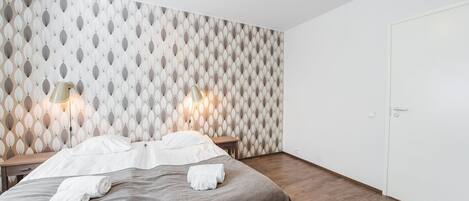 A hotel room with a patterned wallpaper, a bed with white linens, two bedside lamps, and a wooden floor.