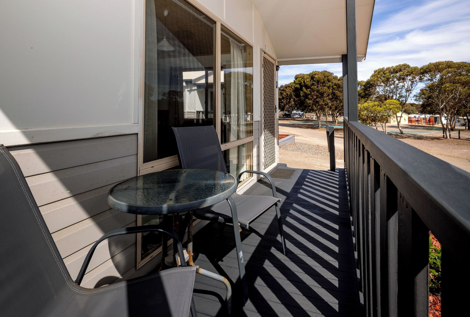 A balcony with a glass table and chairs, a white building, and trees in the background.