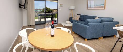 A modern living room with a round wooden table, white chairs, a blue sofa, and a large window offering a view of the outdoors.