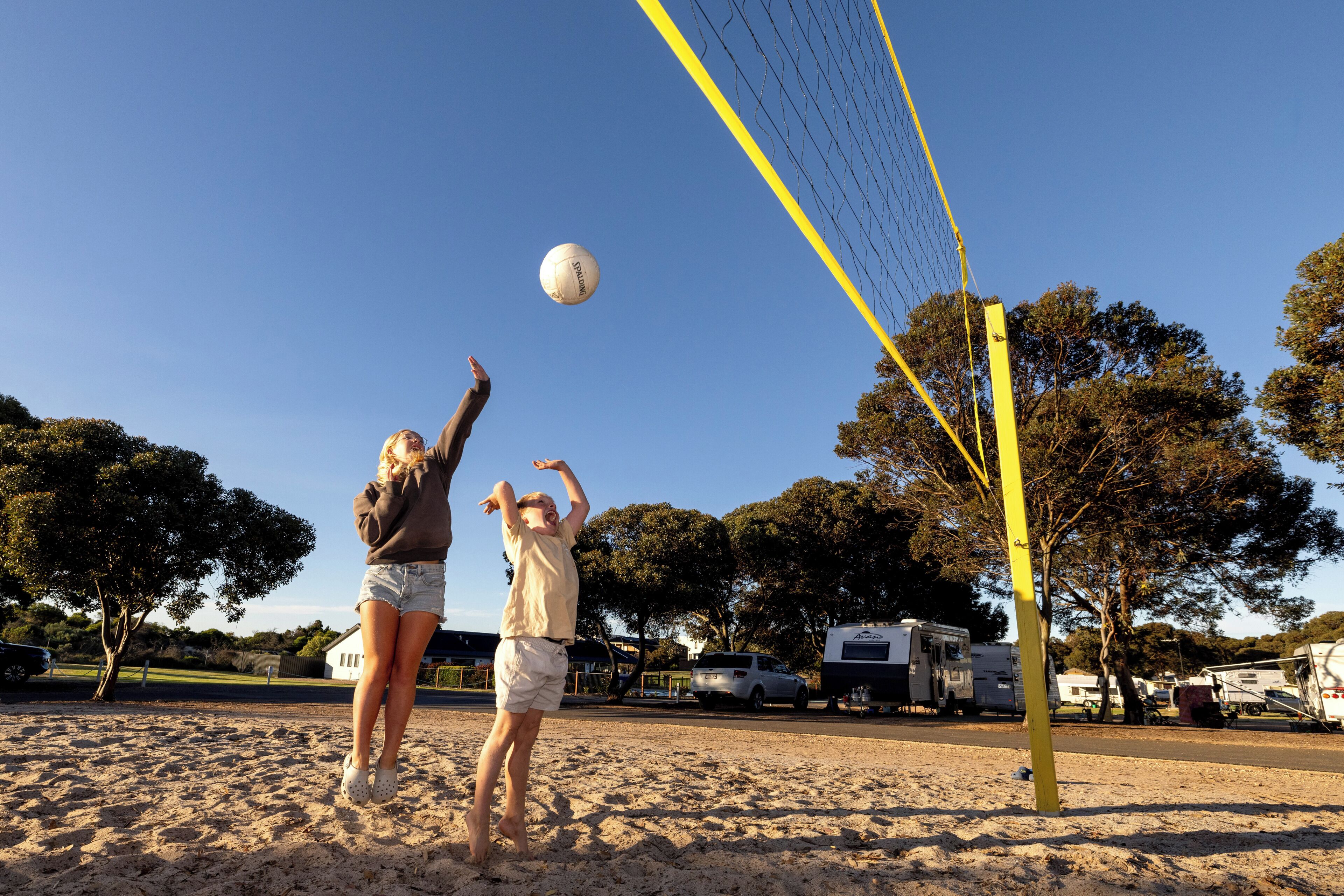 Two people playing volleyball on a sandy court with trees and RVs in the background.