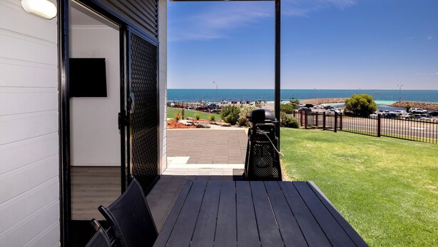 An outdoor patio with a view of the sea, a barbecue grill, and a paved area with cars.