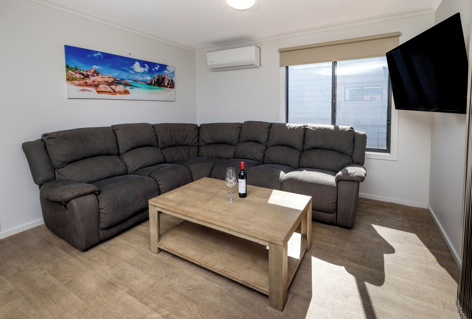 Een moderne woonkamer met een modulaire bank, een houten salontafel, een schilderij van een strandgezicht, een airconditioning, een raam met jaloezieën en een flatscreen televisie aan de muur.
