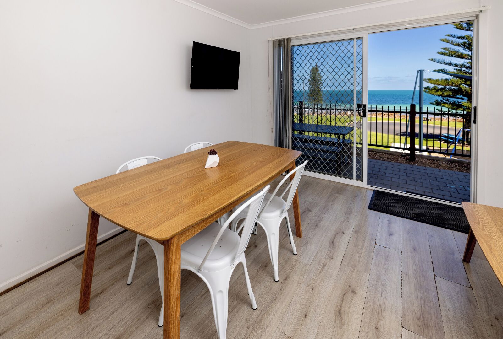 A room with a wooden table, white chairs, a TV mounted on the wall, and a sliding glass door leading to a balcony with a view of the sea.