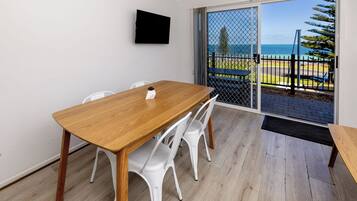 A room with a wooden table, white chairs, a TV mounted on the wall, and a sliding glass door leading to a balcony with a view of the sea.