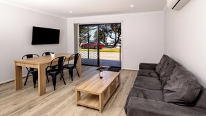 A living room with a wooden table, black chairs, a grey sofa, a flat-screen TV, and a sliding glass door leading to an outdoor area.