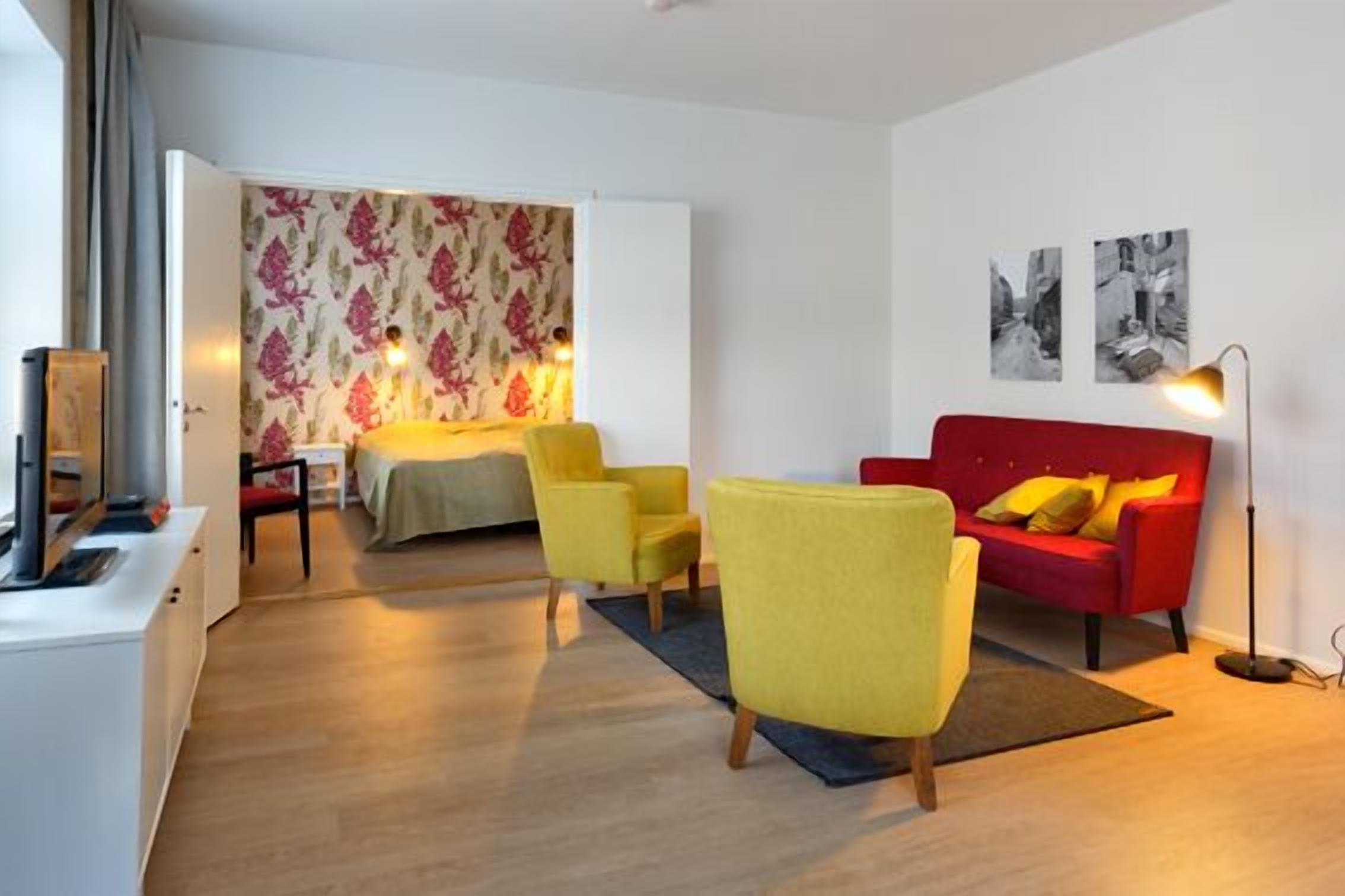 A modern living room with a red sofa, yellow armchairs, and a bed with floral wallpaper.