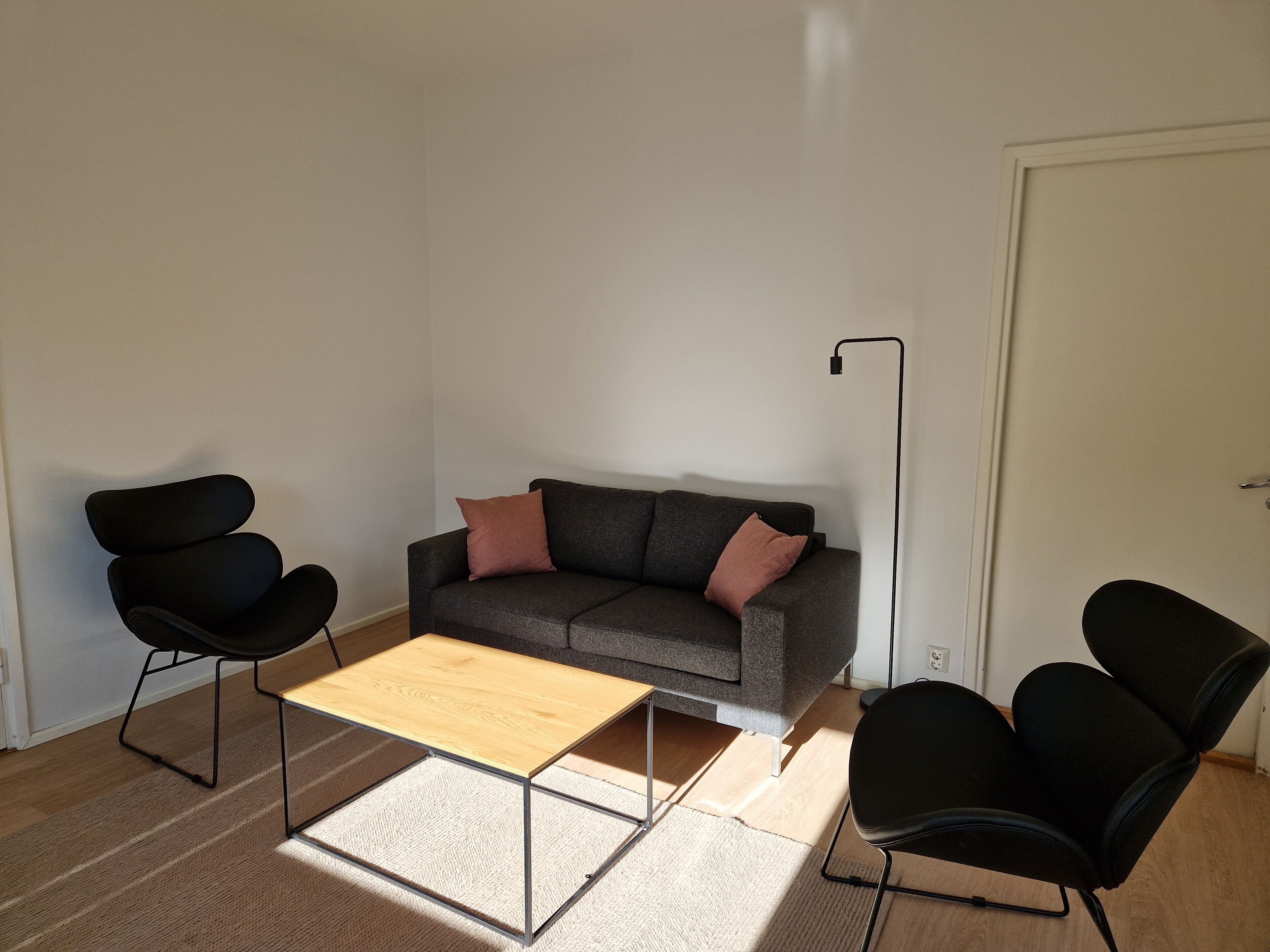 Sala de estar com sofá cinza, duas cadeiras pretas, mesa de centro de madeira e luminária de chão.