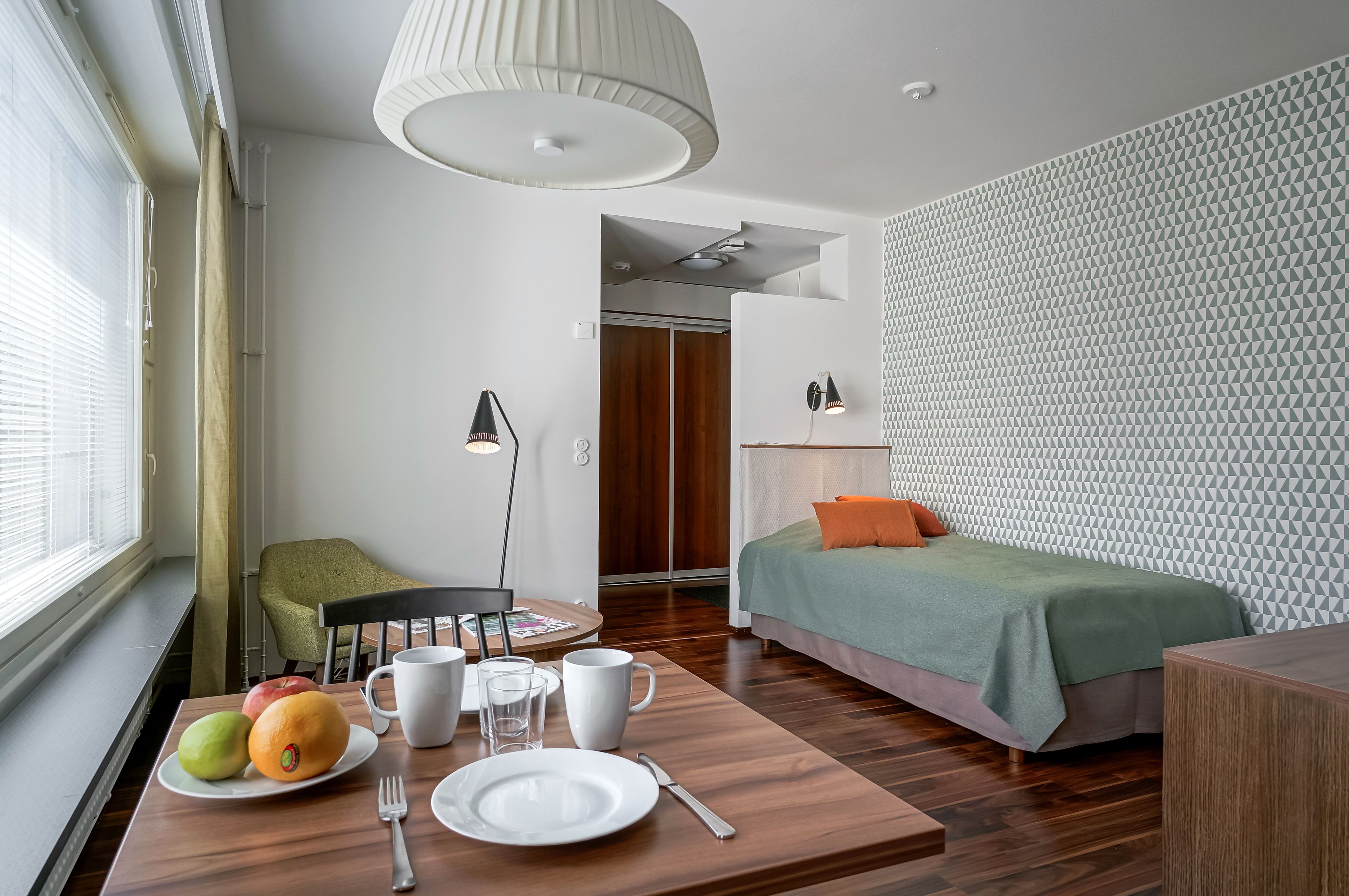 Une chambre d’hôtel moderne avec un plancher en bois, un lit vert, une table à manger avec des fruits et des assiettes, et un mur orné de motifs.