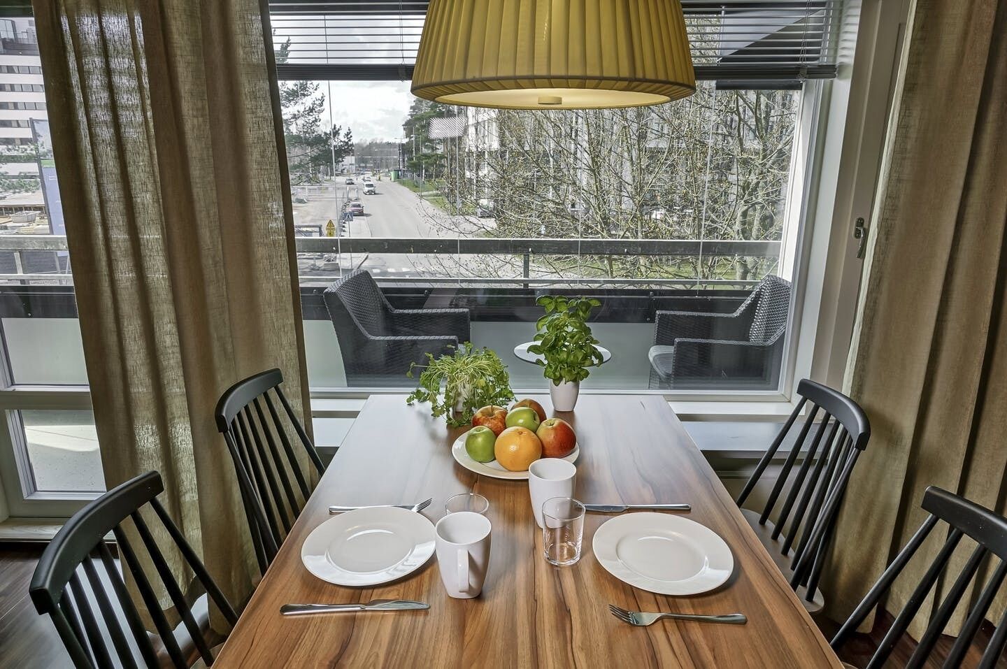 A dining table with plates, cutlery, a glass, and a fruit bowl, set against a window with a view of a street and buildings.