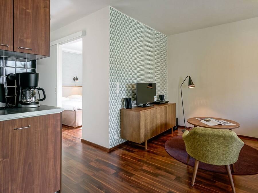 Quarto de hotel moderno com um armário de madeira, uma cafeteira, uma televisão sobre um suporte de madeira, uma cadeira verde e uma pequena mesa redonda com uma revista.