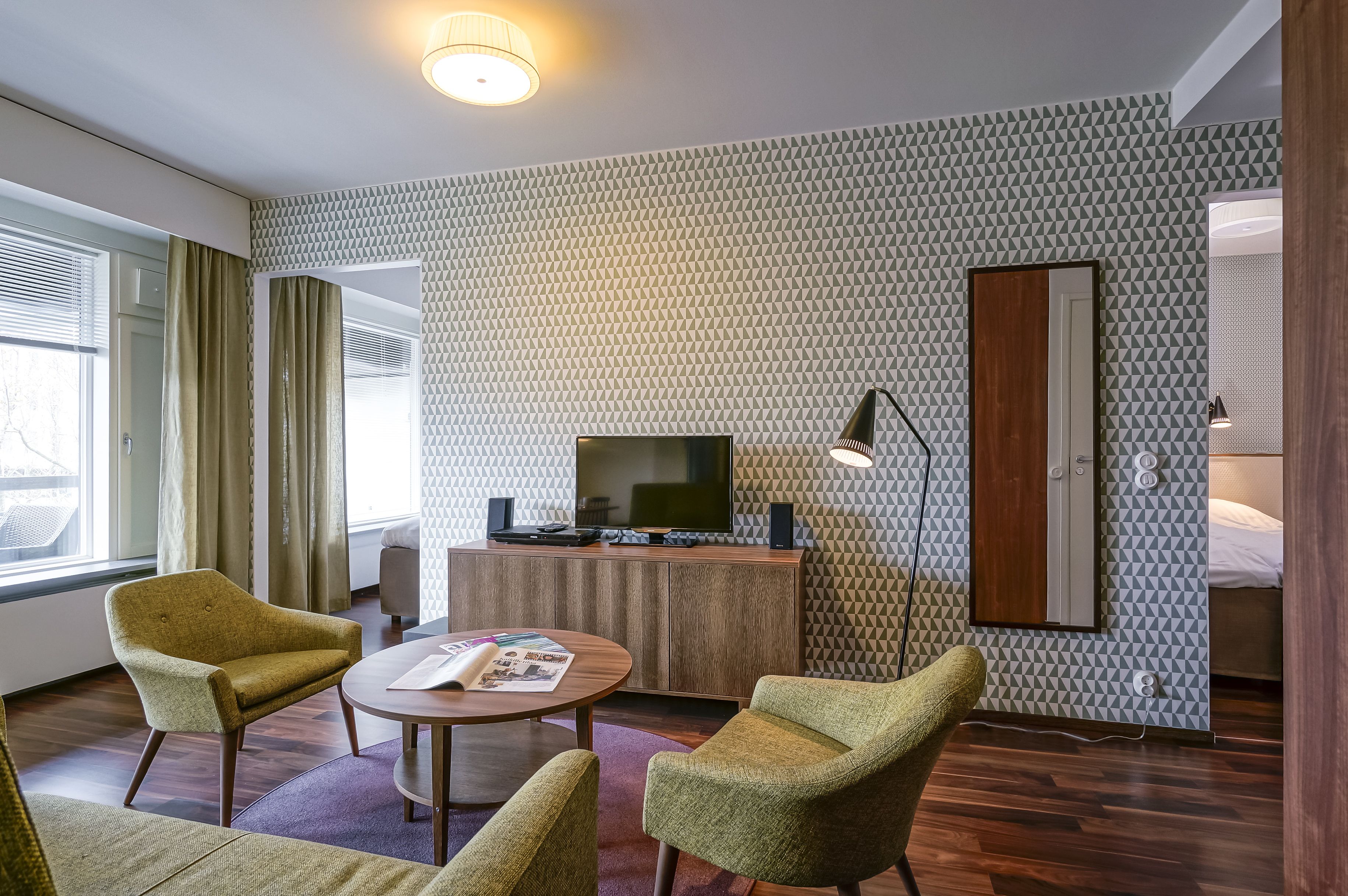 A modern hotel room with a patterned wall, a flat-screen TV, a wooden cabinet, and two green armchairs.