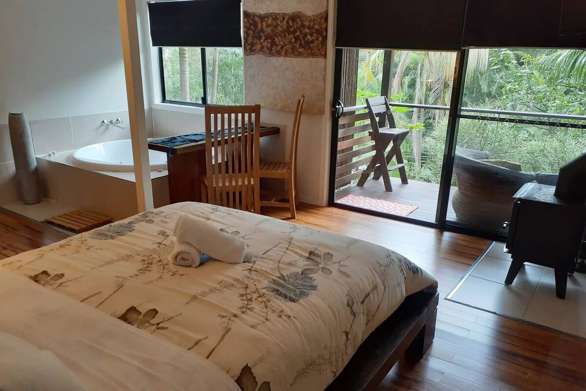 A bedroom with a large bed, a wooden chair, a small table, a bathtub, and a balcony with a view of greenery.