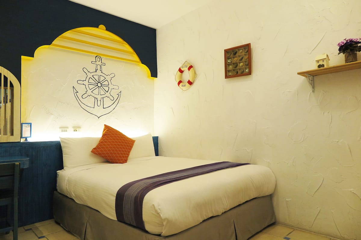 Una cama bien hecha con una colcha blanca y morada, una silla de madera, un salvavidas colgado en la pared y una pequeña estantería con una planta.