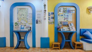 Una habitación con dos escritorios de marco azul, cada uno con un taburete pequeño, un tablón de anuncios con varios papeles y un mapa, y un banco acolchado azul.