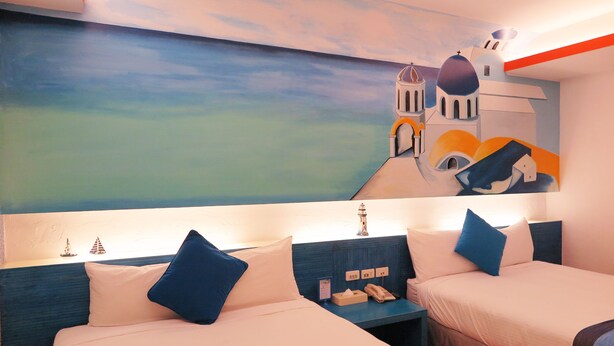 Habitación de hotel con dos camas, un mural de paisaje costero y una mesita de noche con lámpara y teléfono.