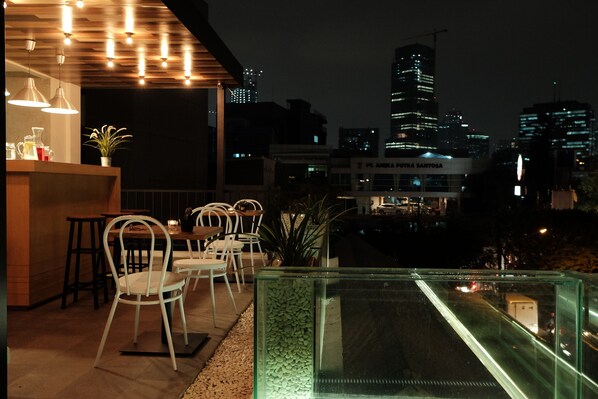 A rooftop bar with a glass enclosure, white chairs, and a cityscape view at night. - Flat06 Tendean (Jakarta)