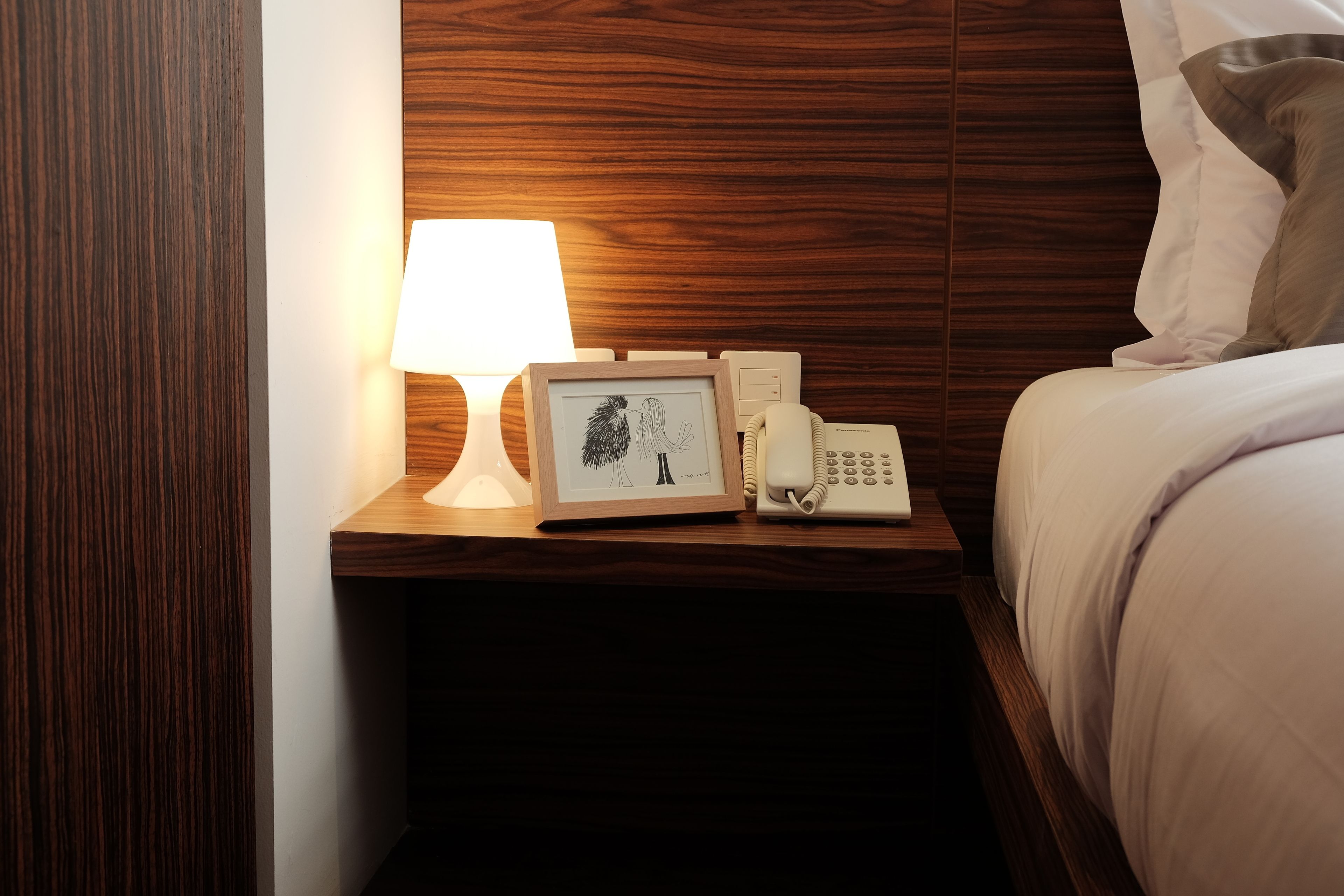Una cama bien hecha con ropa blanca, una lámpara de noche, un cuadro enmarcado y un teléfono sobre una mesita de madera.