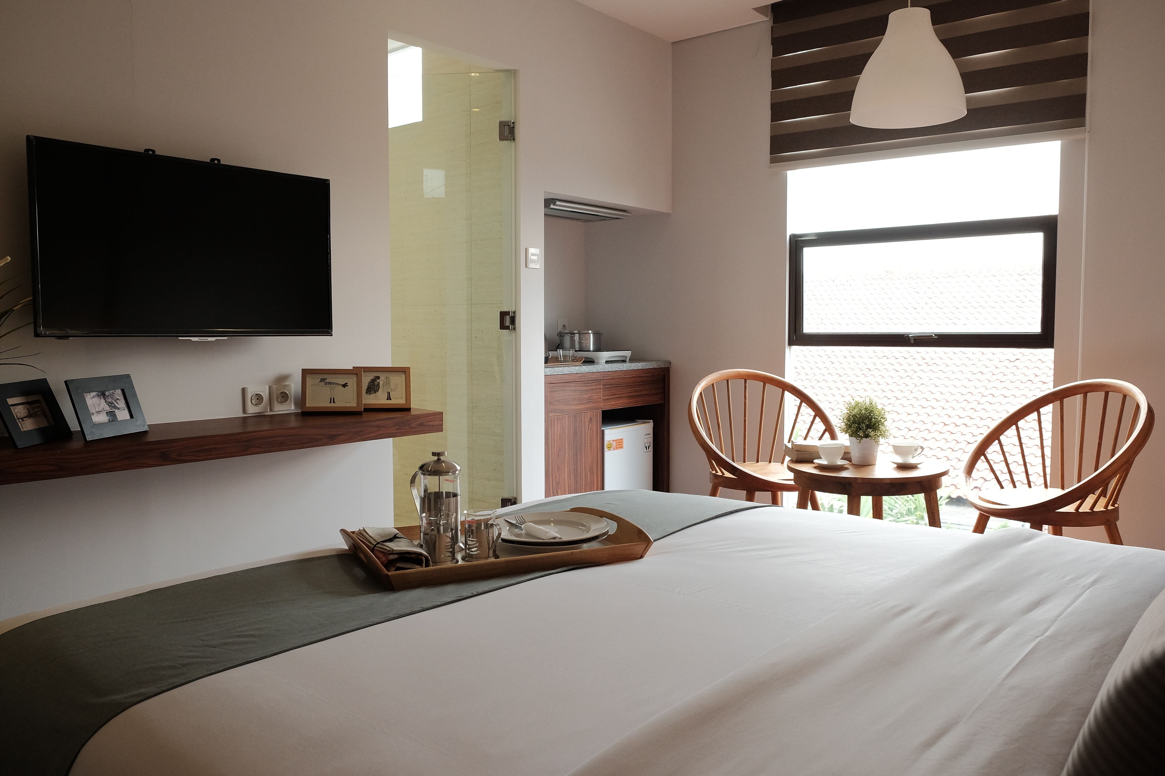 Quarto de hotel moderno com uma cama grande, televisão na parede, área de refeições com duas cadeiras, uma mesinha com uma planta e uma janela com vista.