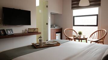 Quarto de hotel moderno com uma cama grande, televisão na parede, área de refeições com duas cadeiras, uma mesinha com uma planta e uma janela com vista.