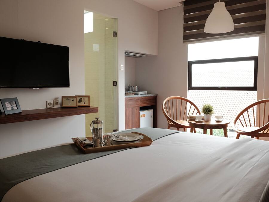 Quarto de hotel moderno com uma cama grande, televisão na parede, área de refeições com duas cadeiras, uma mesinha com uma planta e uma janela com vista.