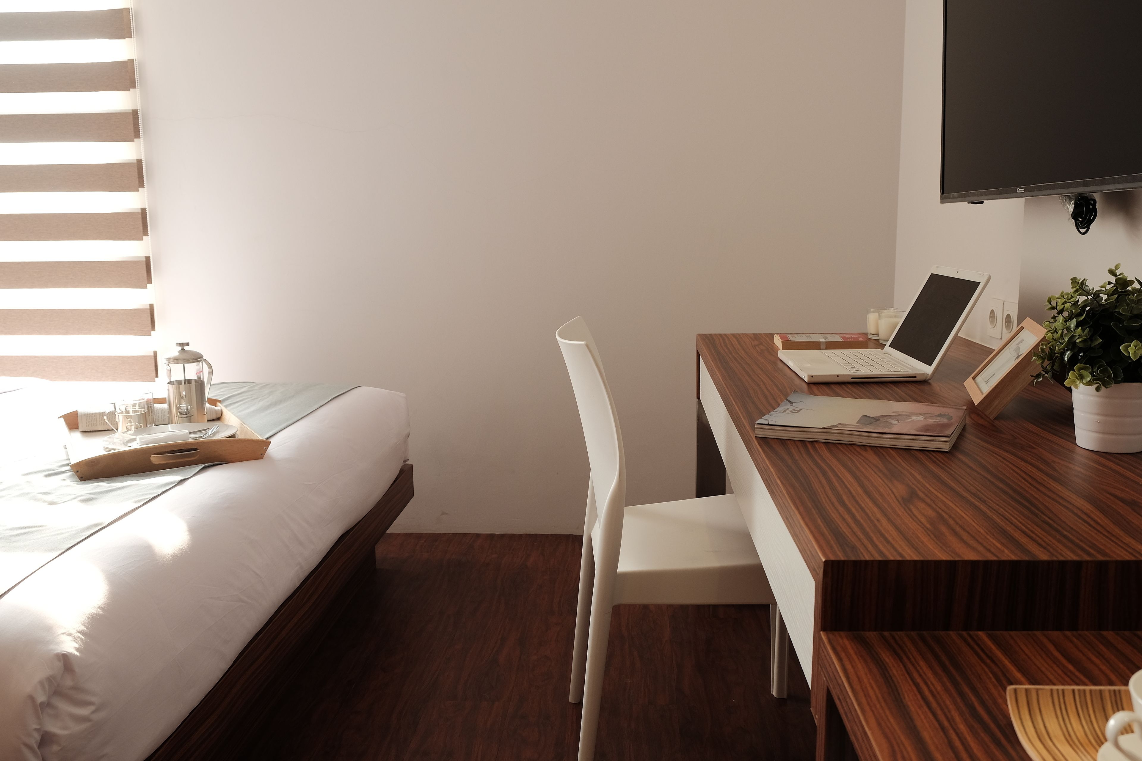 Une chambre d’hôtel avec un bureau en bois, un ordinateur portable, une chaise blanche, un lit et un téléviseur fixé au mur.