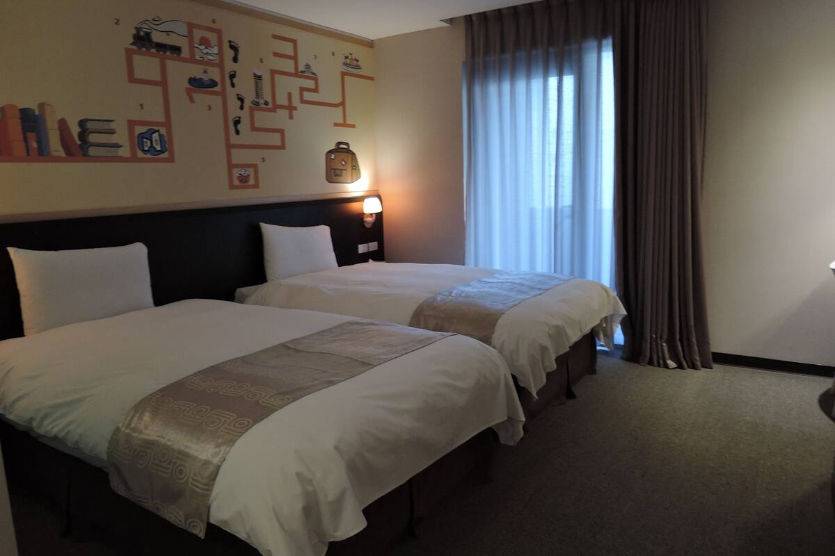 A hotel room with two beds, a headboard with a light, a wall with a maze-like design, and a window with curtains.