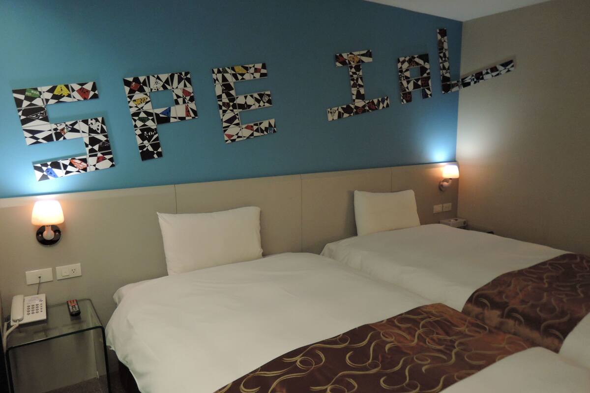 A hotel room with two beds, a blue wall with mosaic letters spelling "SPEAK," a glass nightstand with a phone, and a brown patterned throw blanket.