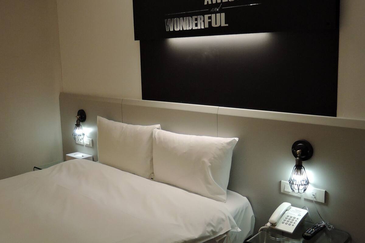 A hotel room with a bed, bedside lamps, a wall-mounted phone, and a motivational quote on the wall.