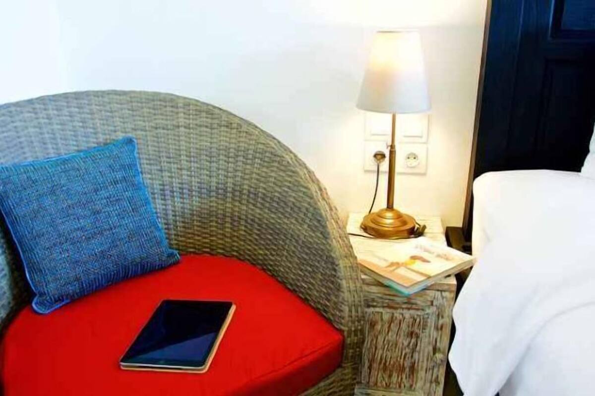A cozy room with a curved grey sofa, a red cushion, a blue pillow, and a bedside table with a lamp and a book.