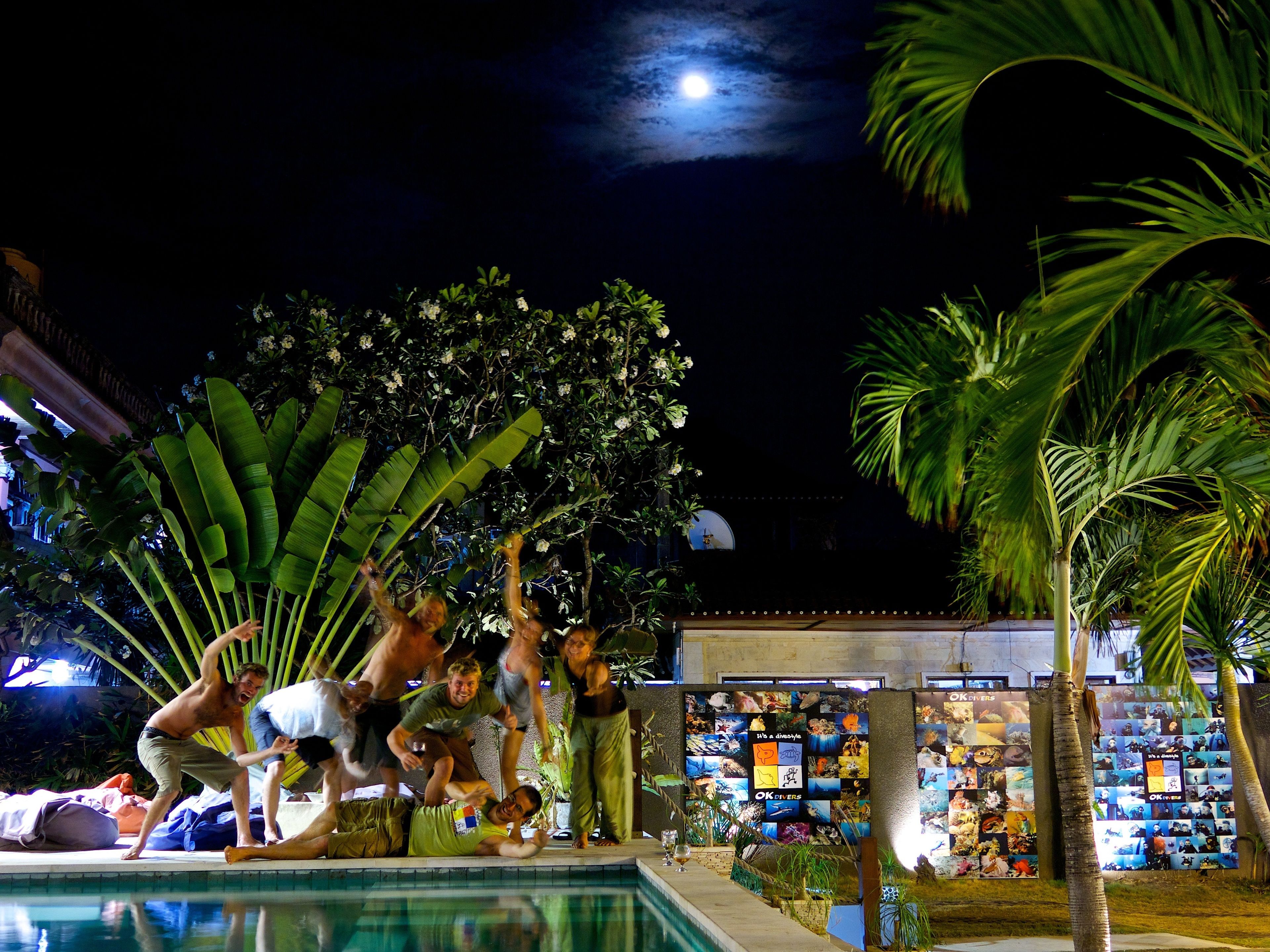 Zona de piscina con gente disfrutando, rodeada de exuberante vegetación y palmeras.