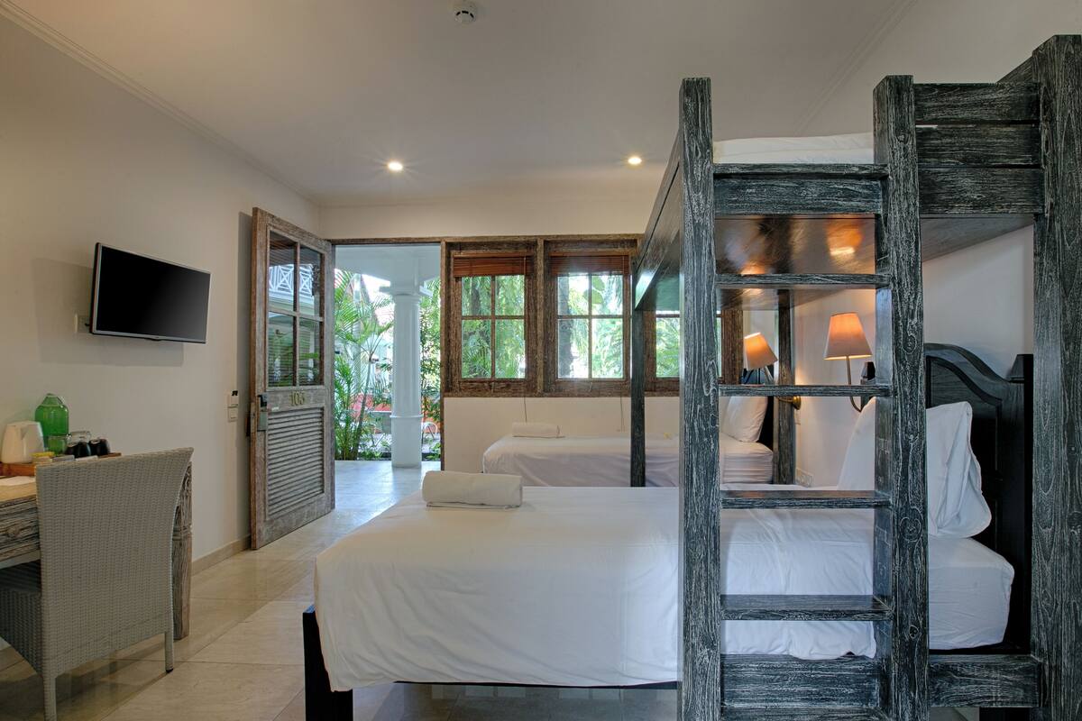 A bedroom with two beds, a desk, a television, and a view of greenery through large windows.