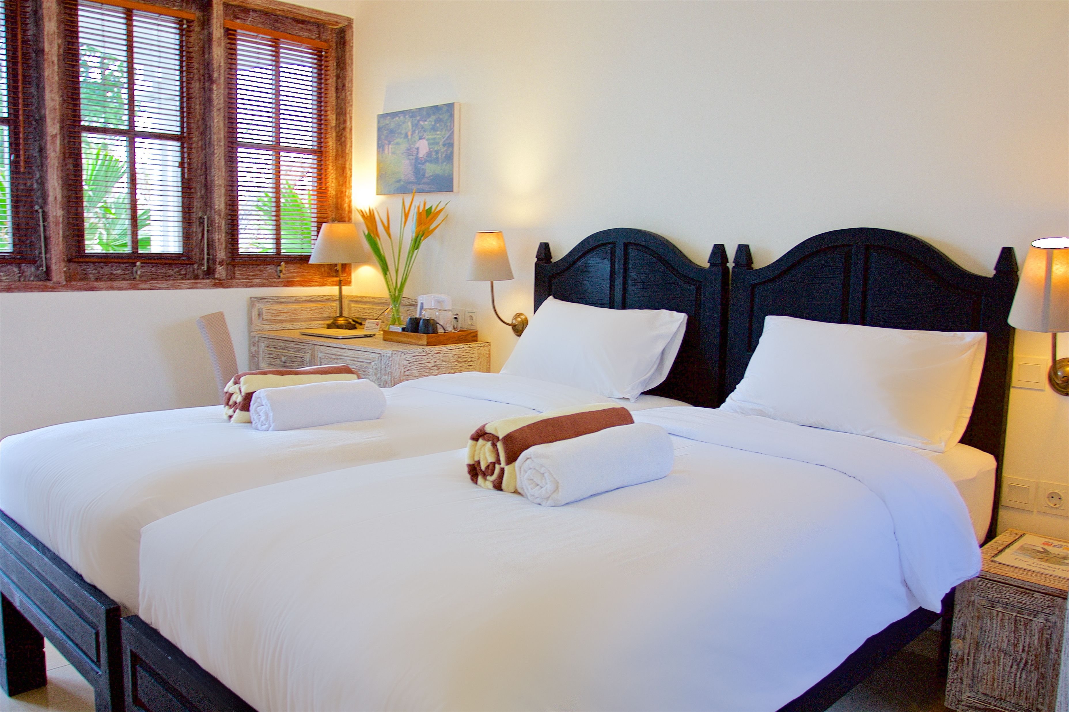 A double bed with white linens, two bedside tables with lamps, a window with wooden shutters, and a painting on the wall.