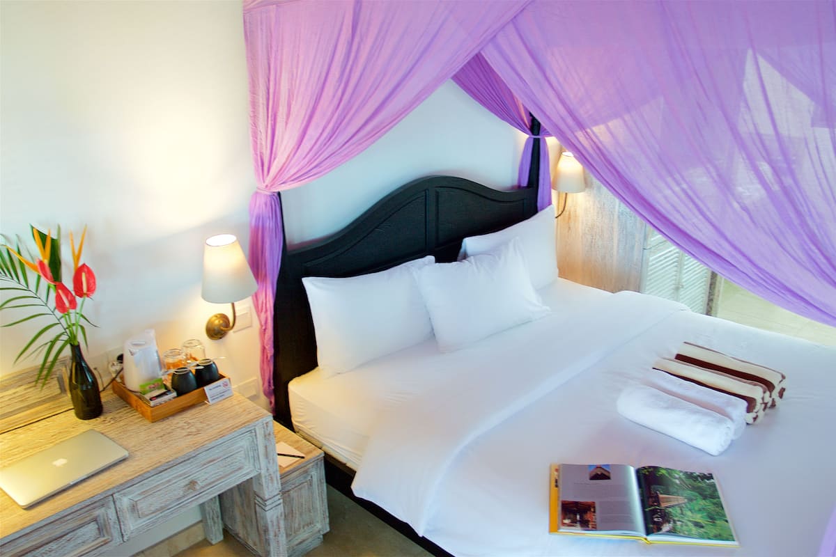 A neatly made bed with white linens and a canopy, a bedside table with a lamp, a laptop, and a tray with books and a vase.