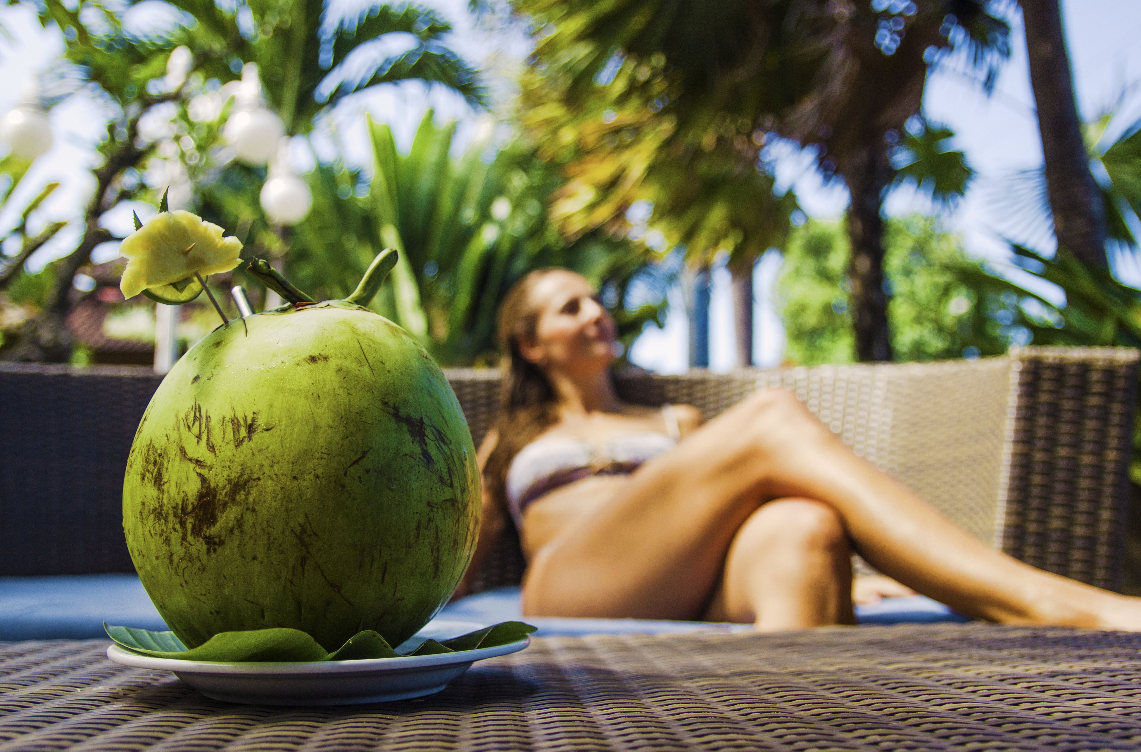 A green coconut on a plate with a leaf, a wicker chair, and a person reclining in the background.