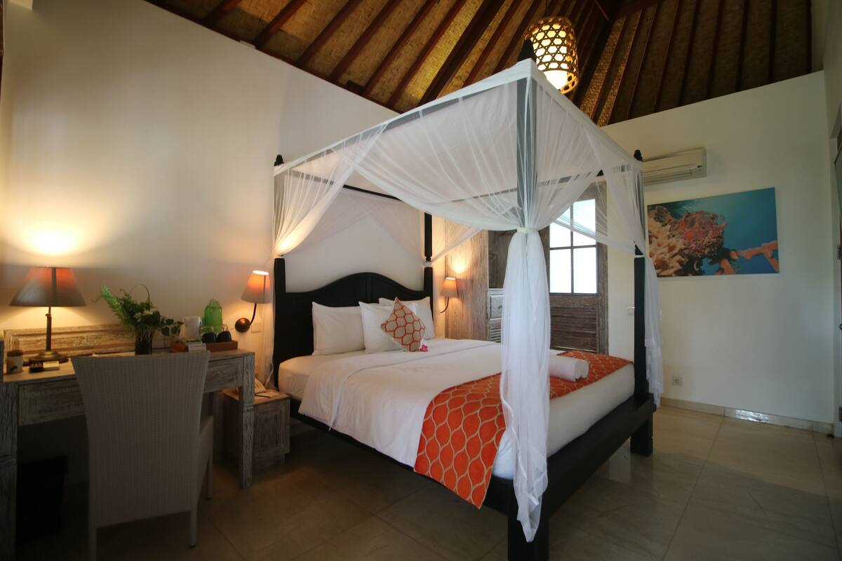 A four-poster bed with a canopy, a desk with a lamp, and a chair in a room with wooden beams on the ceiling.