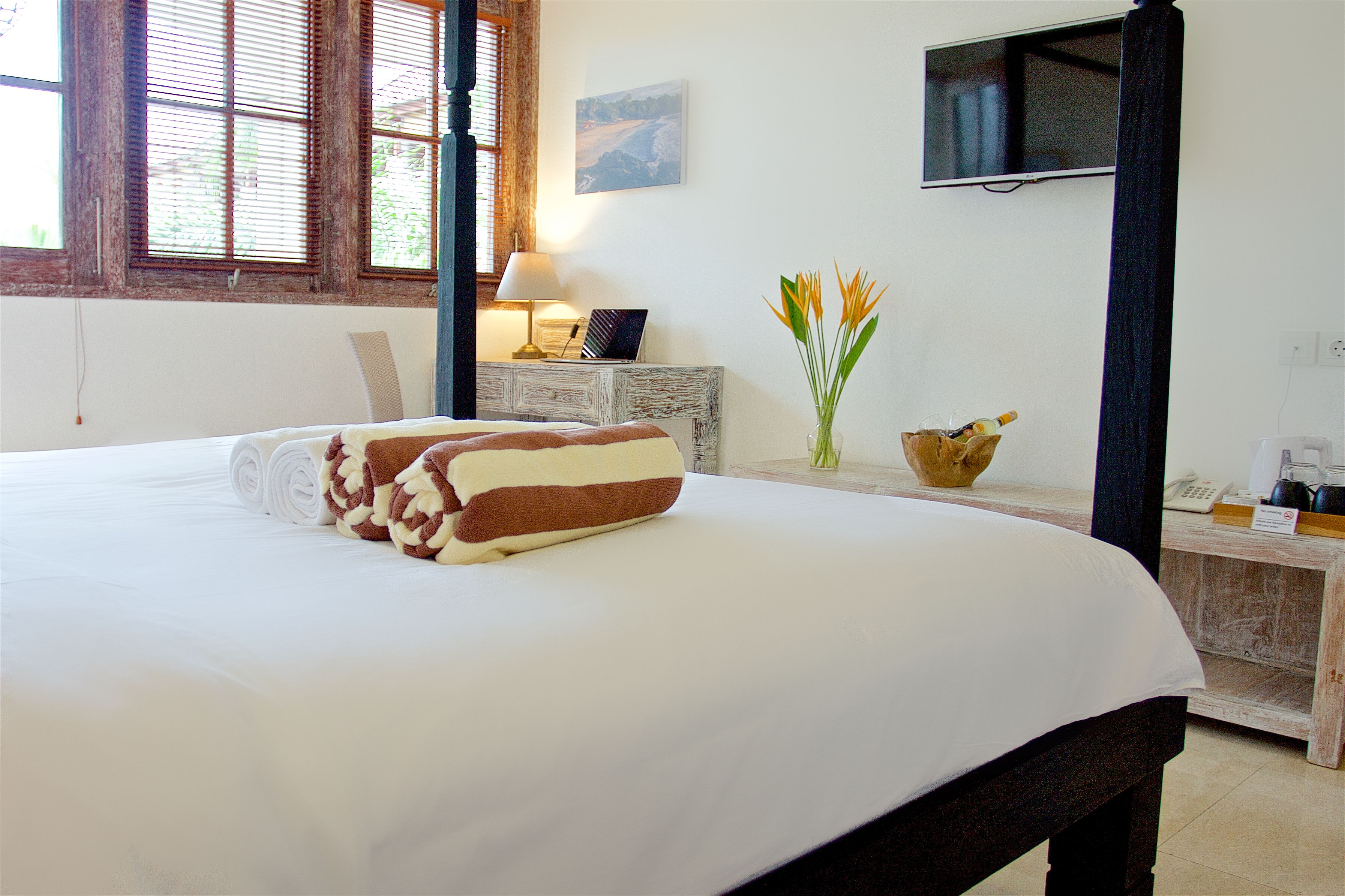 A neatly made bed with rolled towels, a bedside table with a lamp, a vase with flowers, and a TV mounted on the wall.