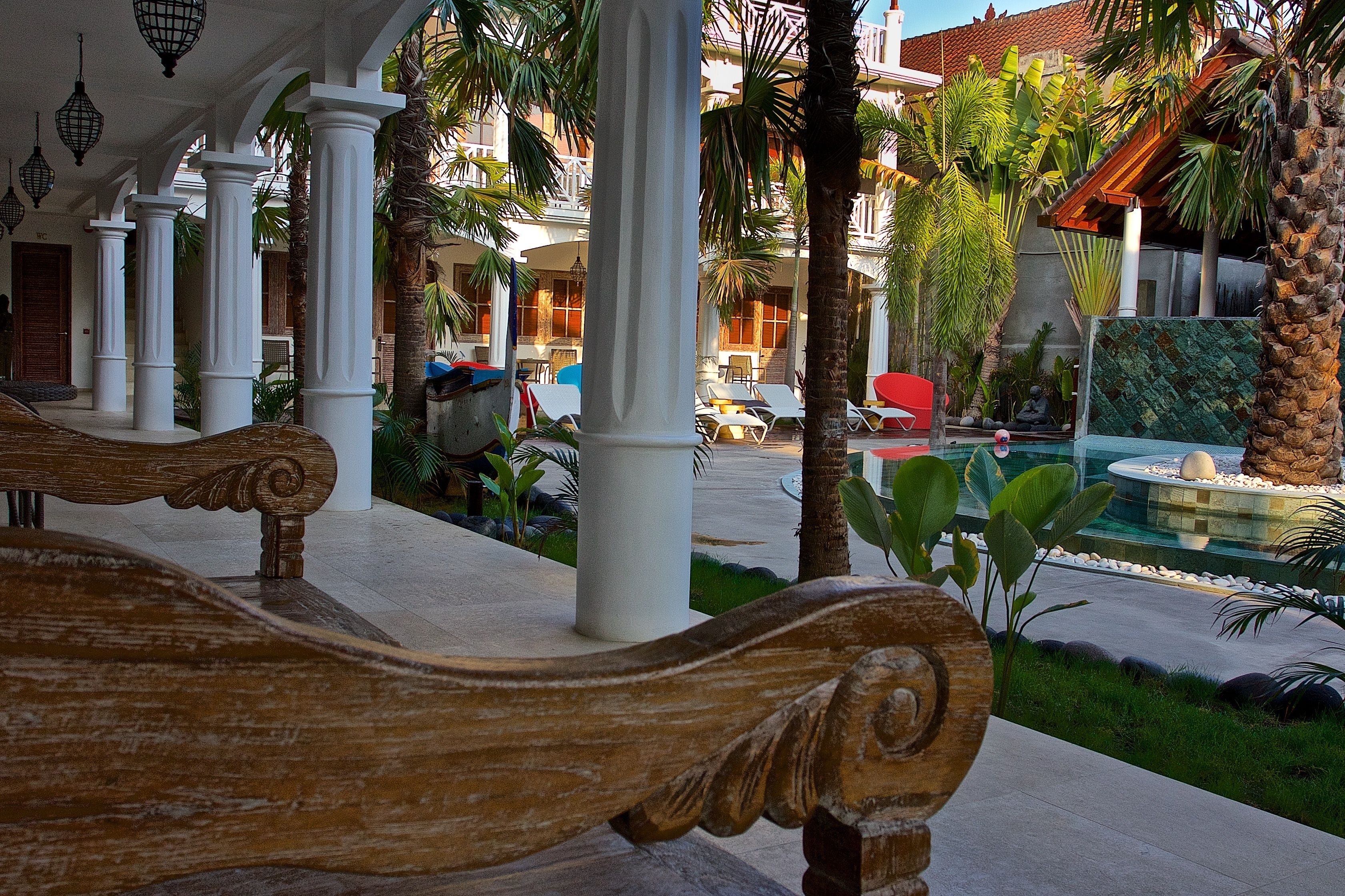 Un espacio al aire libre techado con columnas blancas, un banco de madera, sillones de descanso y una pequeña piscina con una fuente de piedra.