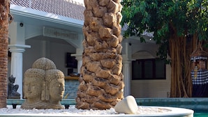 A stone Buddha statue, a rock formation, and a pool area with a palm tree and a building in the background. - OK Divers Resort & Spa (Padangbai)