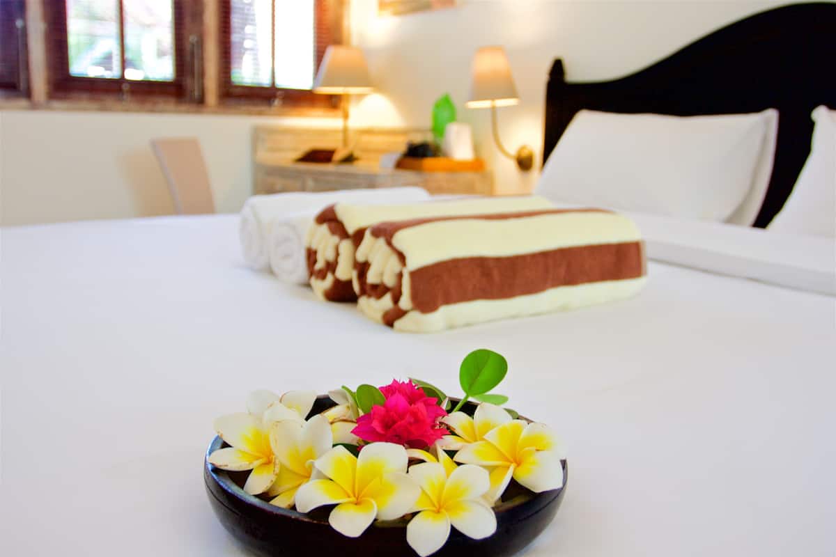 A neatly made bed with a pillow and a rolled towel, a bowl of flowers, and a lamp on a bedside table.