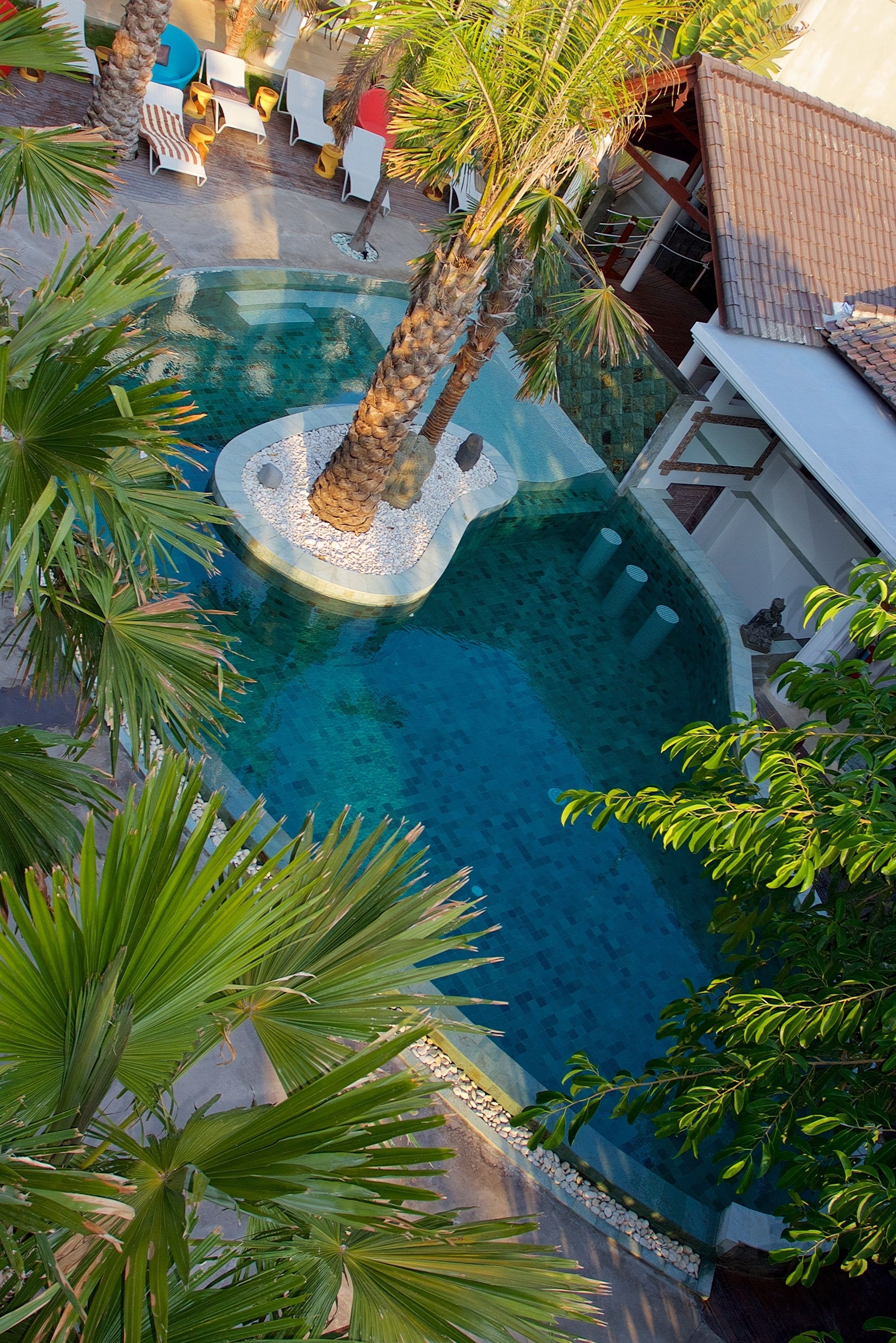 A swimming pool with a palm tree centerpiece, surrounded by lounge chairs and a tiled roof structure.