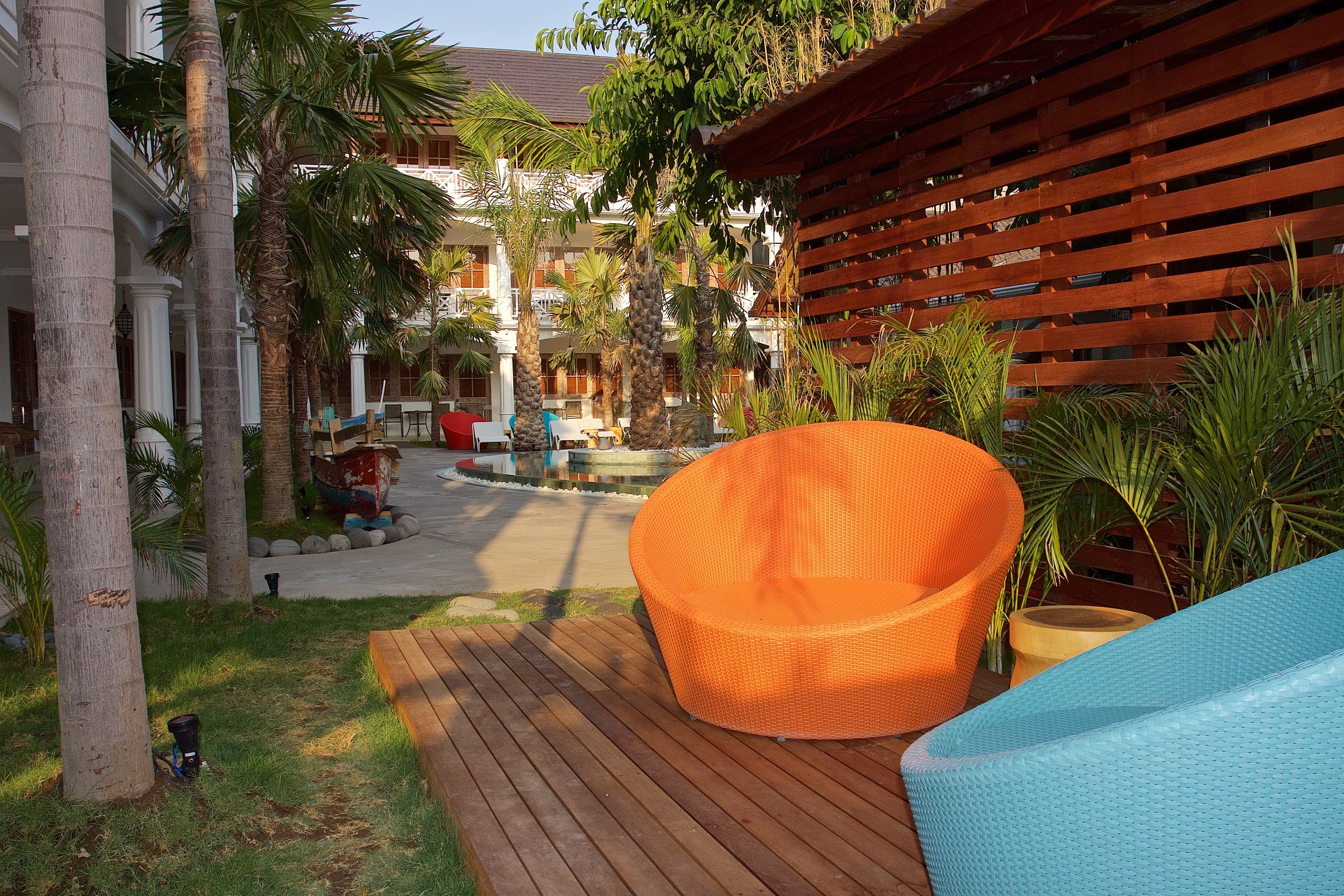 Une terrasse en bois avec un fauteuil en osier, des palmiers et, au fond, un bâtiment doté de balcons.