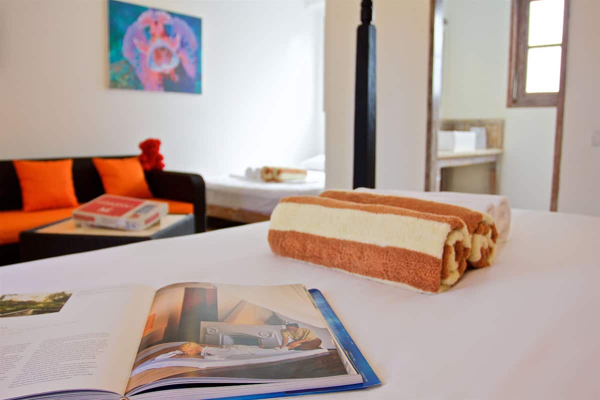 A hotel room with a bed, a book, a towel, a small table with a box, and a colorful painting on the wall.