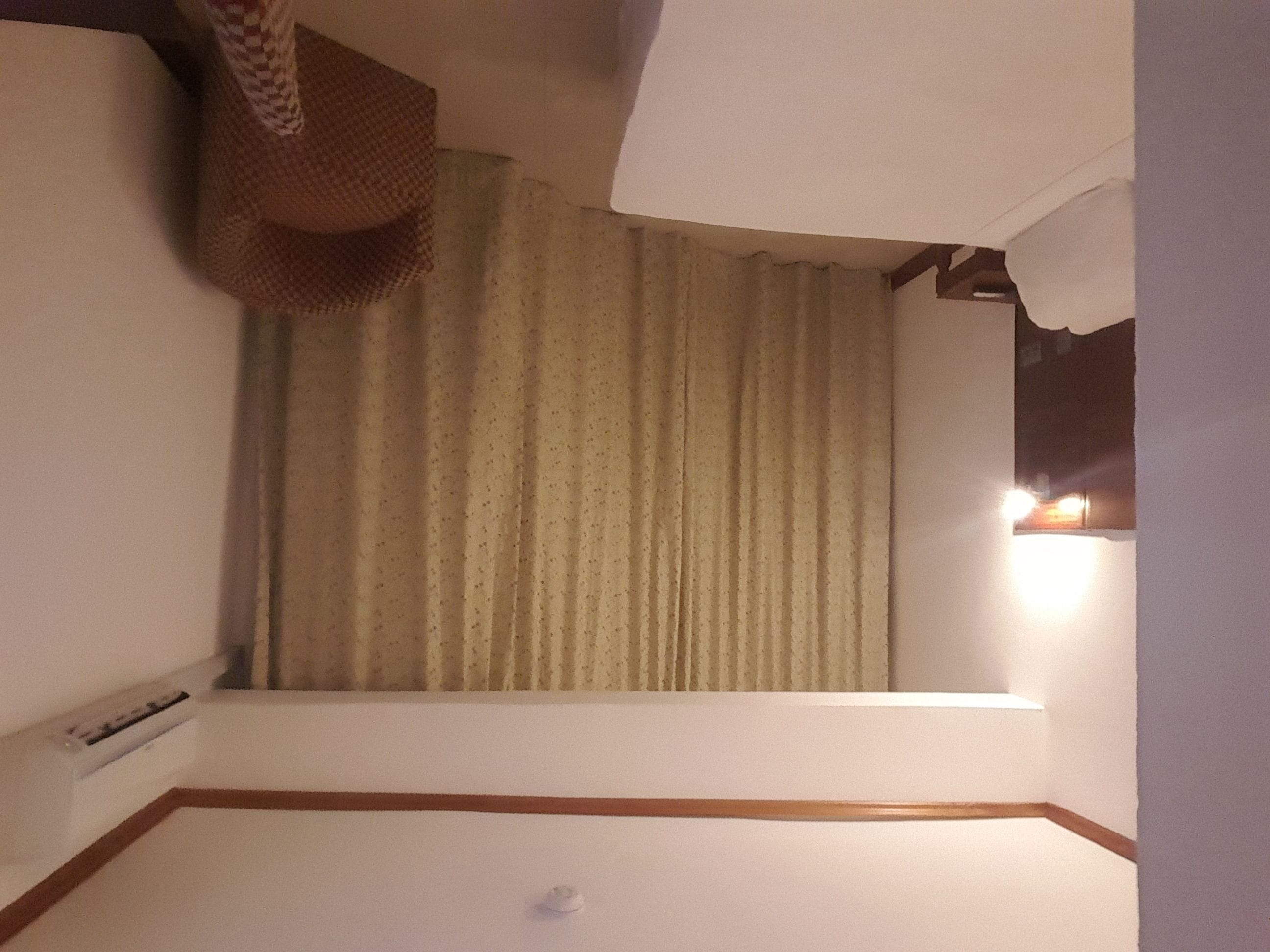 A hotel room with a large window, a curtain, a wall-mounted air conditioning unit, and a wooden trim along the baseboard.